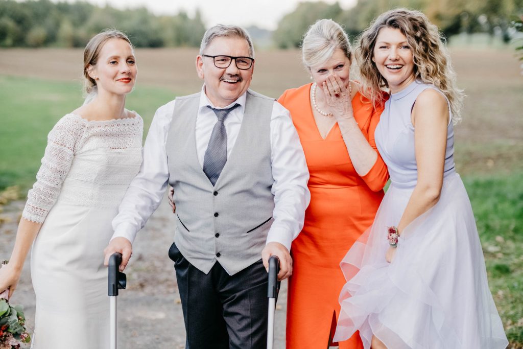 Braut mit Schwester und Eltern lachend bei einem Gruppenfoto im Freien, festlich gekleidet.