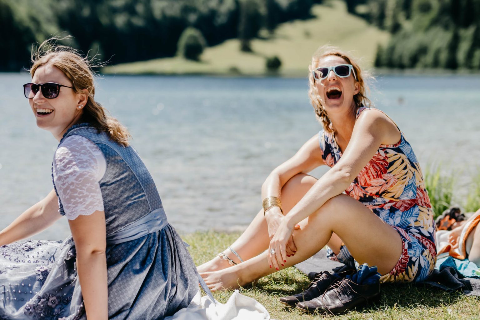 Zwei Hochzeitsgäste im Dirndl und Kleid sitzen lachend auf der Wiese am Alatsee