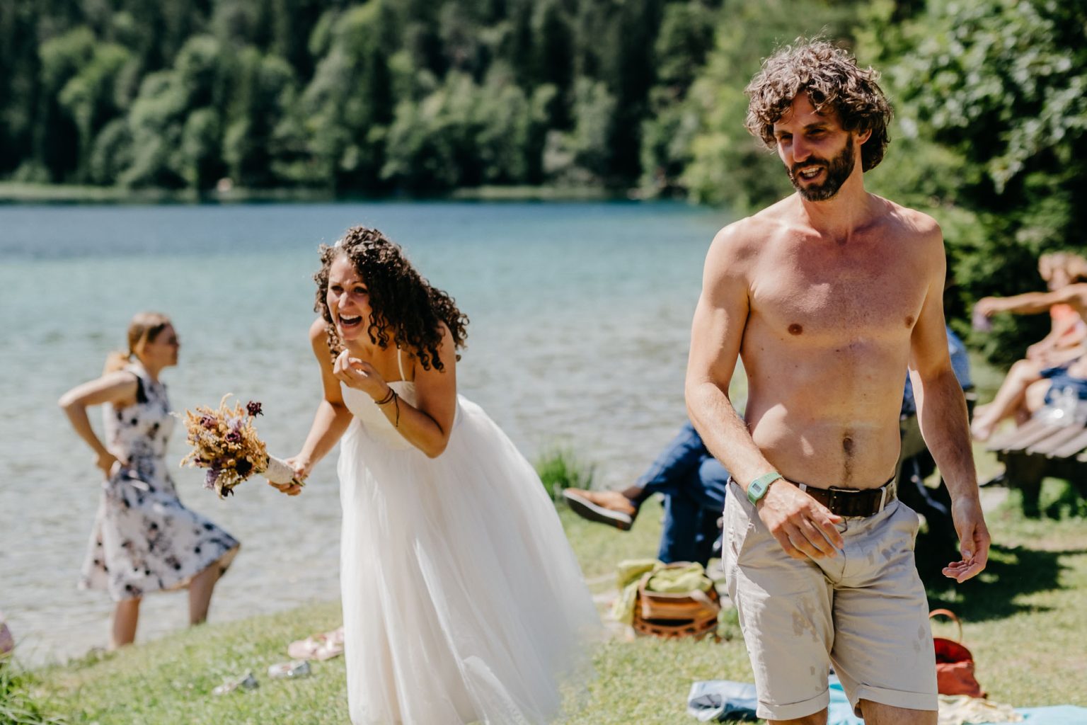 Braut lacht beim Sektempfang am See – im Vordergrund ein männlicher Gast oben ohne