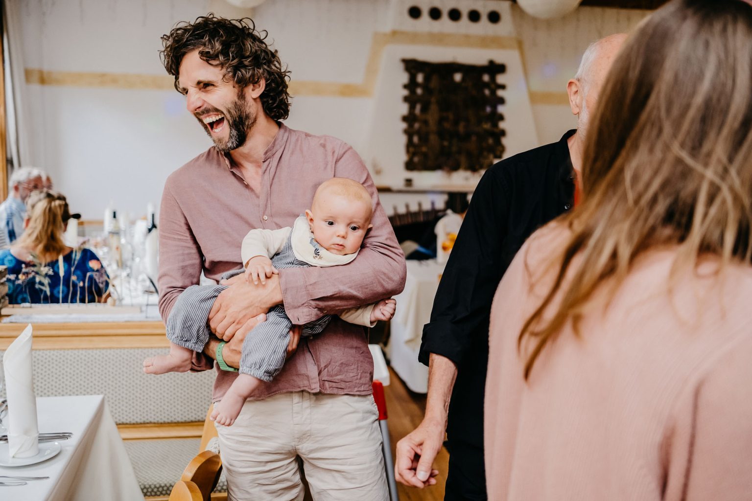 Freund lacht herzlich mit Baby auf dem Arm bei Hochzeit im Allgäu