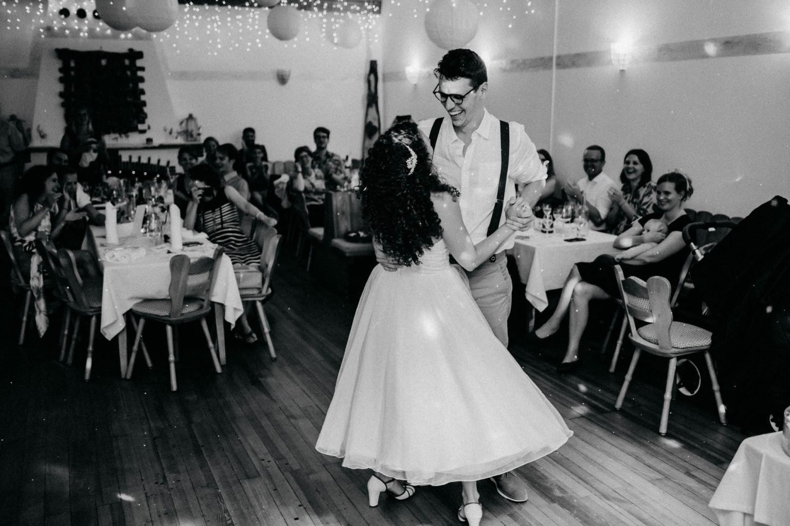 Hochzeitstanz mit lachendem Brautpaar und Hochzeitsgästen im Hintergrund, Schwarz-Weiß