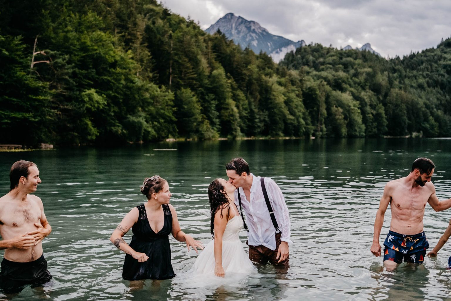 Kuss des Brautpaars im Wasser des Alatsees bei Füssen, Hochzeitsgäste um sie herum.
