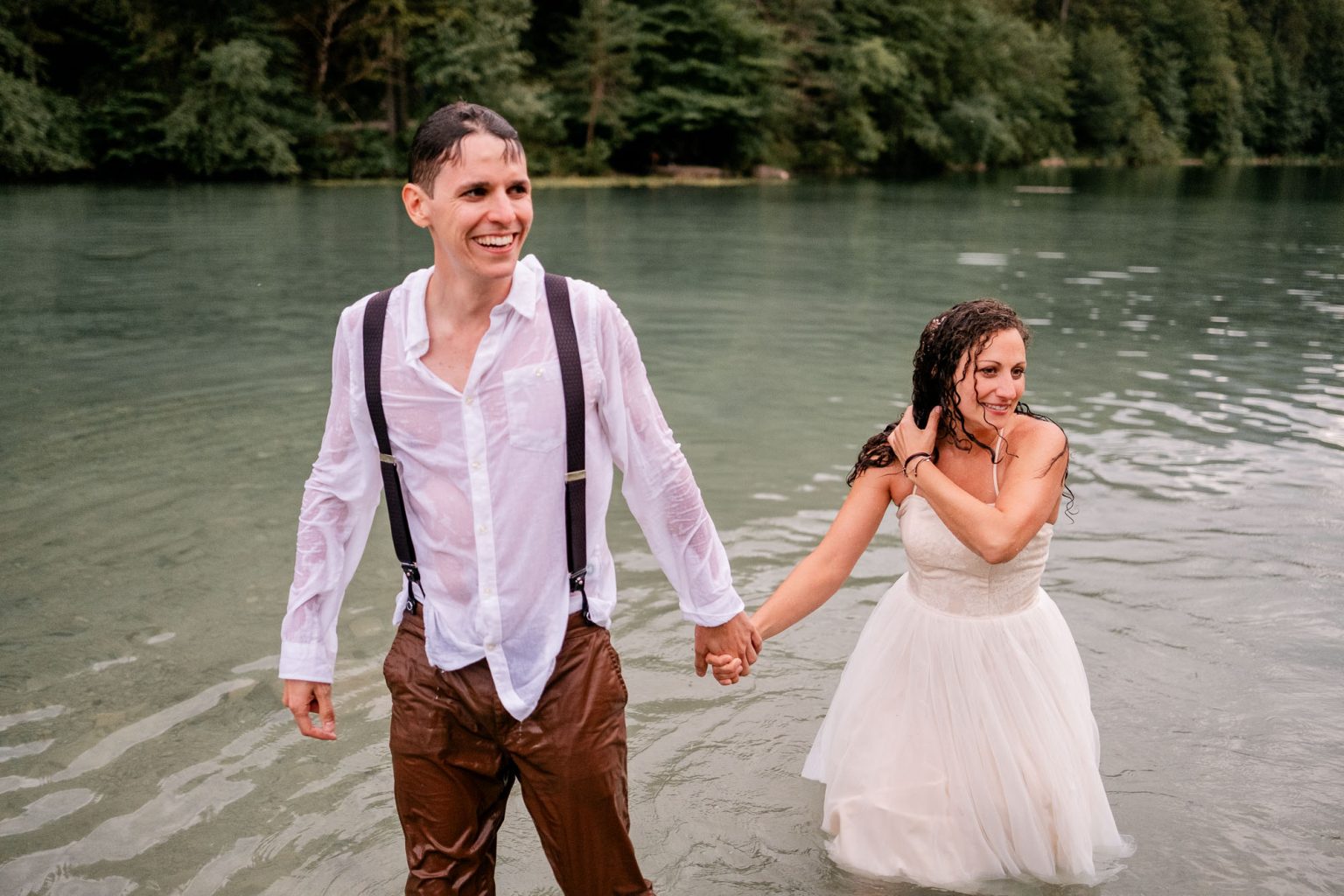 Hochzeitspaar hält sich an den Händen, schaut nach rechts, See und Berge im Hintergrund.